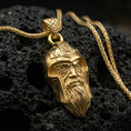 Load image into Gallery viewer, Gold plated Odin pendant necklace on textured volcanic rock, premium close-up with warm reflective shine across the Viking helmet, stern expression, and sculpted beard lines.
