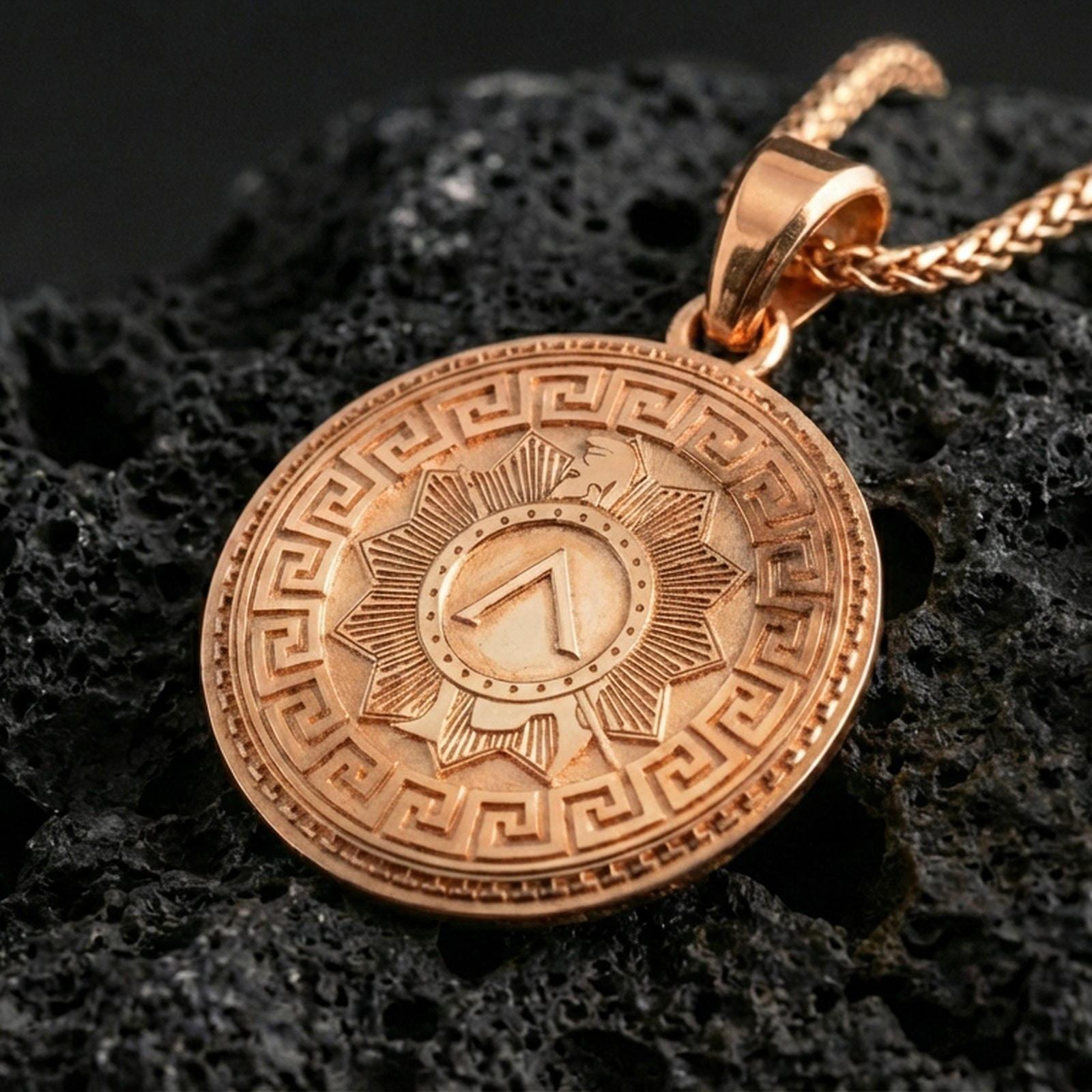 Rose gold plated Spartan shield medallion pendant necklace on textured volcanic rock, centered studio-style close-up showcasing soft rosy glow and detailed ancient Greek patterning.