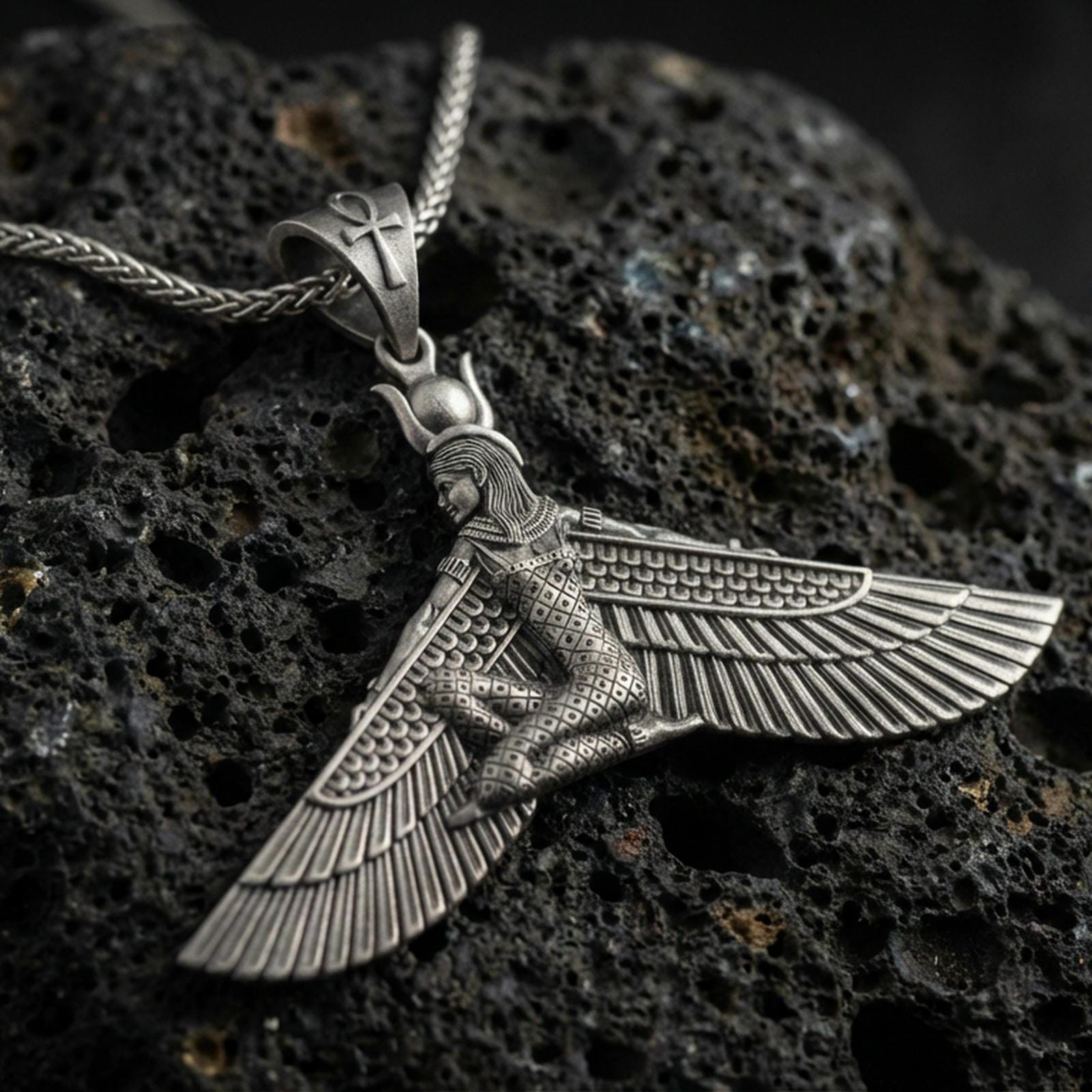 Oxidized winged goddess ankh pendant necklace on textured volcanic rock, centered close-up highlighting the spread wings, ankh bale detail, and deep antique shadowed contrast.