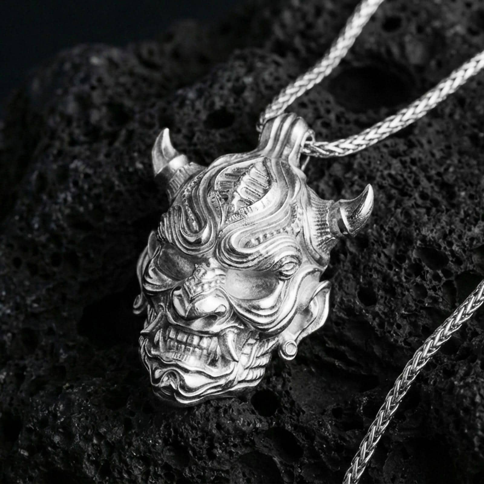 Polished silver oni mask pendant necklace on textured volcanic rock, crisp macro product photo emphasizing clean edges, sharp facial lines, and bright reflective highlights on the sculpted details.