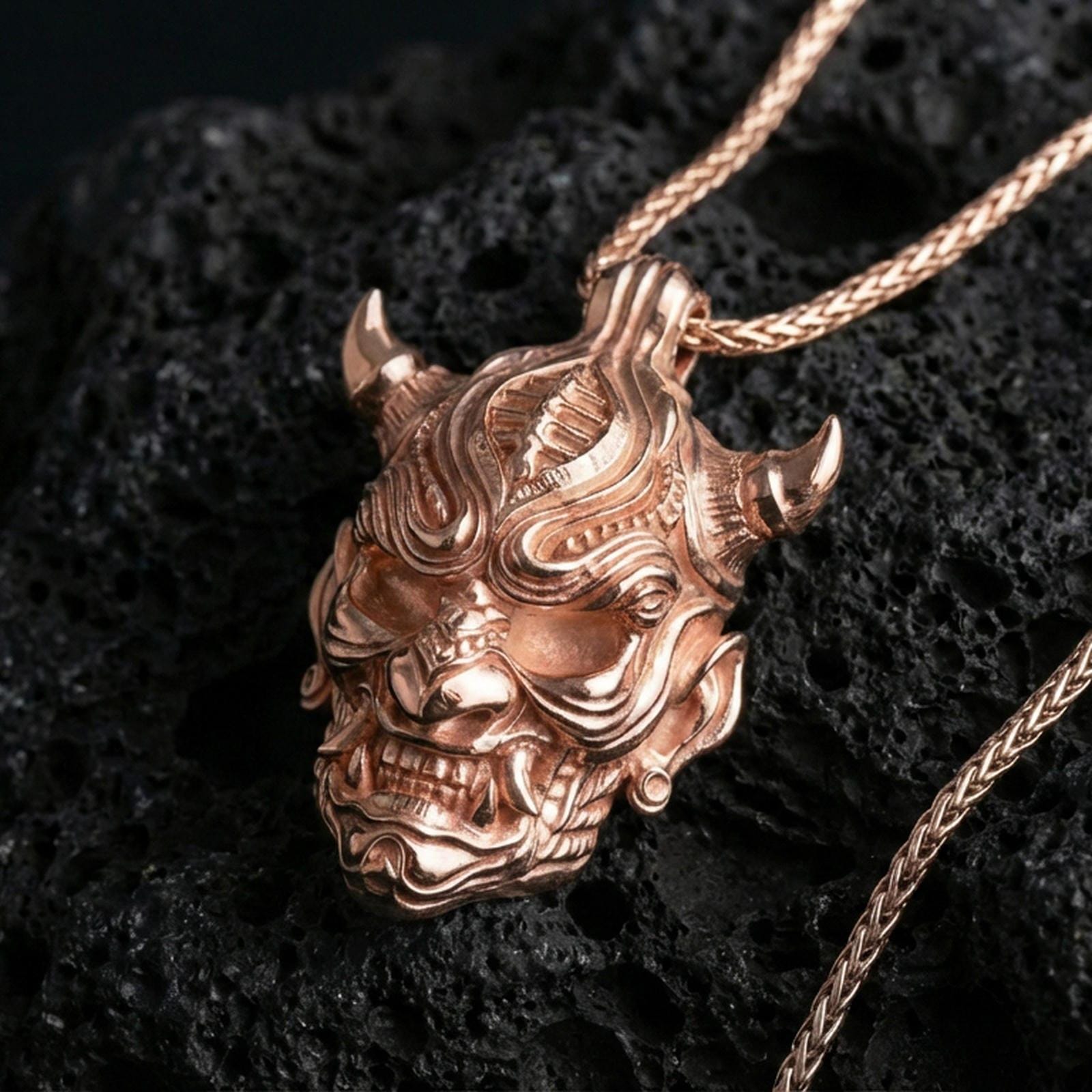 Rose gold plated oni mask pendant necklace on textured volcanic rock, centered studio-style close-up showcasing soft rosy glow and intricate samurai-inspired demon mask detailing.