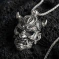 Load image into Gallery viewer, Oxidized oni mask pendant necklace on textured volcanic rock, centered close-up highlighting the horned demon face, sharp fangs, and deep antique shadowed contrast.
