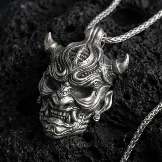 Oxidized oni mask pendant necklace on textured volcanic rock, centered close-up highlighting the horned demon face, sharp fangs, and deep antique shadowed contrast.