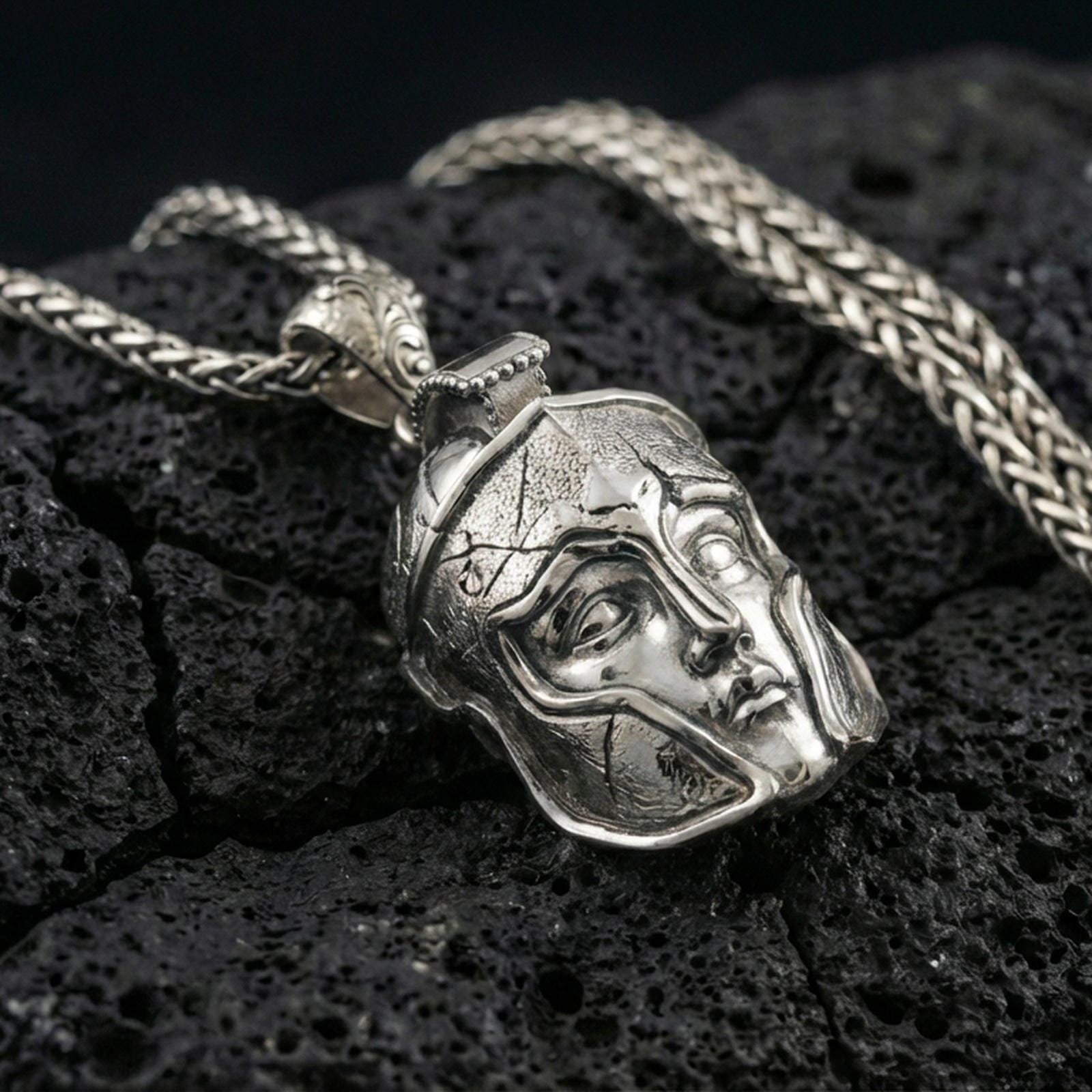 Polished silver Spartan helmet pendant on porous volcanic stone, close-up focusing on the sculpted facial features, sharp contours, and clean reflective edges