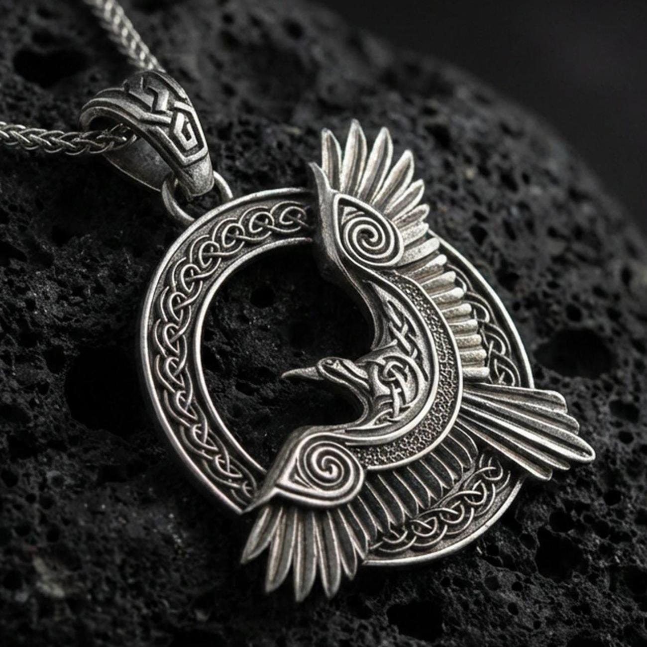 Oxidized Celtic raven knotwork pendant necklace on textured volcanic rock, centered close-up highlighting the winged raven silhouette, spiral accents, and deep antique contrast in the braided frame.