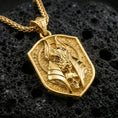 Load image into Gallery viewer, Gold plated Anubis pendant necklace on textured volcanic rock, premium close-up with warm reflective shine across the Anubis head, layered armor-like forms, and raised border.

