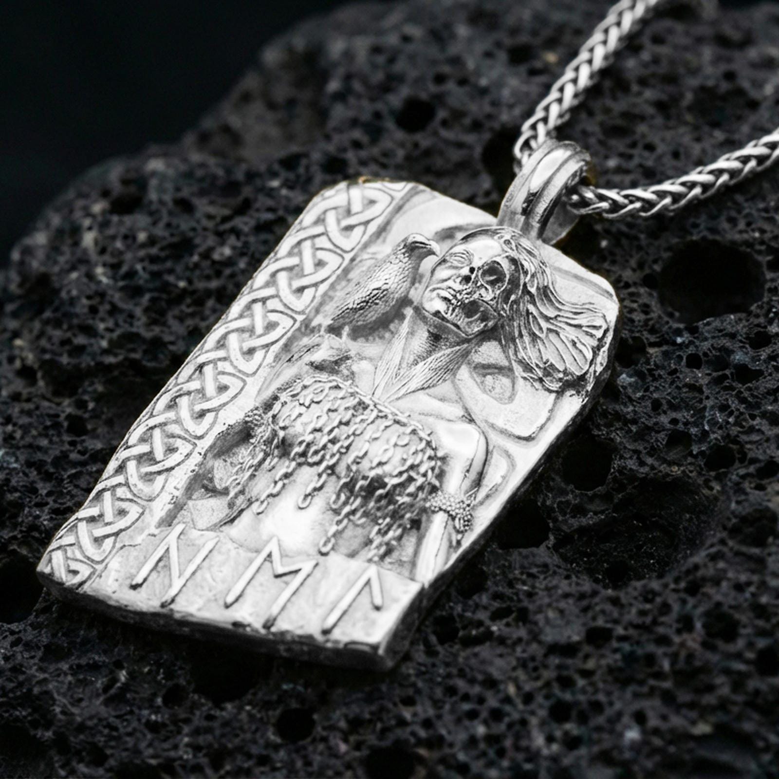Polished silver Hel Norse goddess pendant necklace on textured volcanic rock, crisp macro product photo emphasizing clean edges, sharp facial relief, and detailed textures on the hair, raven, and chain-like armor.