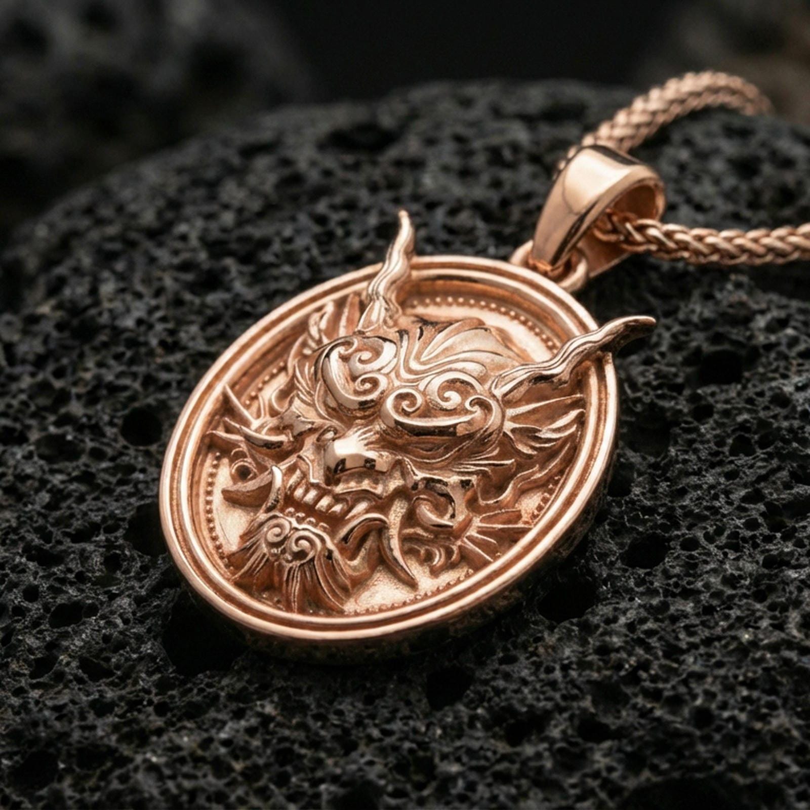 Rose gold plated demon mask medallion pendant necklace on textured volcanic rock, centered studio-style close-up showcasing soft rosy glow and intricate mask-and-fang detailing with a refined finish.