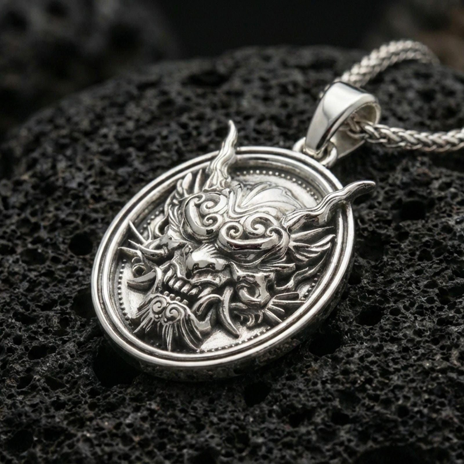 Polished silver demon mask medallion pendant necklace on textured volcanic rock, crisp macro product photo emphasizing clean edges, sharp relief lines, and bright reflective highlights on the sculpted details.
