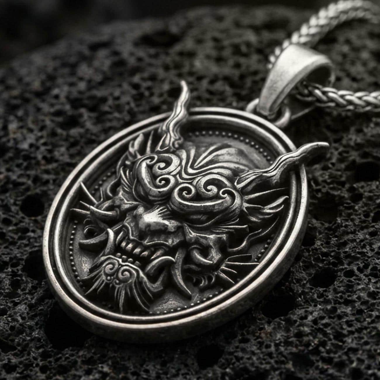 Oxidized demon mask medallion pendant necklace on textured volcanic rock, centered close-up highlighting the horned oni-style face, sharp fangs, and deep antique shadowed contrast.