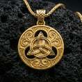 Load image into Gallery viewer, Gold plated Celtic triquetra pendant necklace on textured volcanic rock, premium close-up with warm reflective shine across the Trinity knot, decorative flourishes, and raised border detailing.
