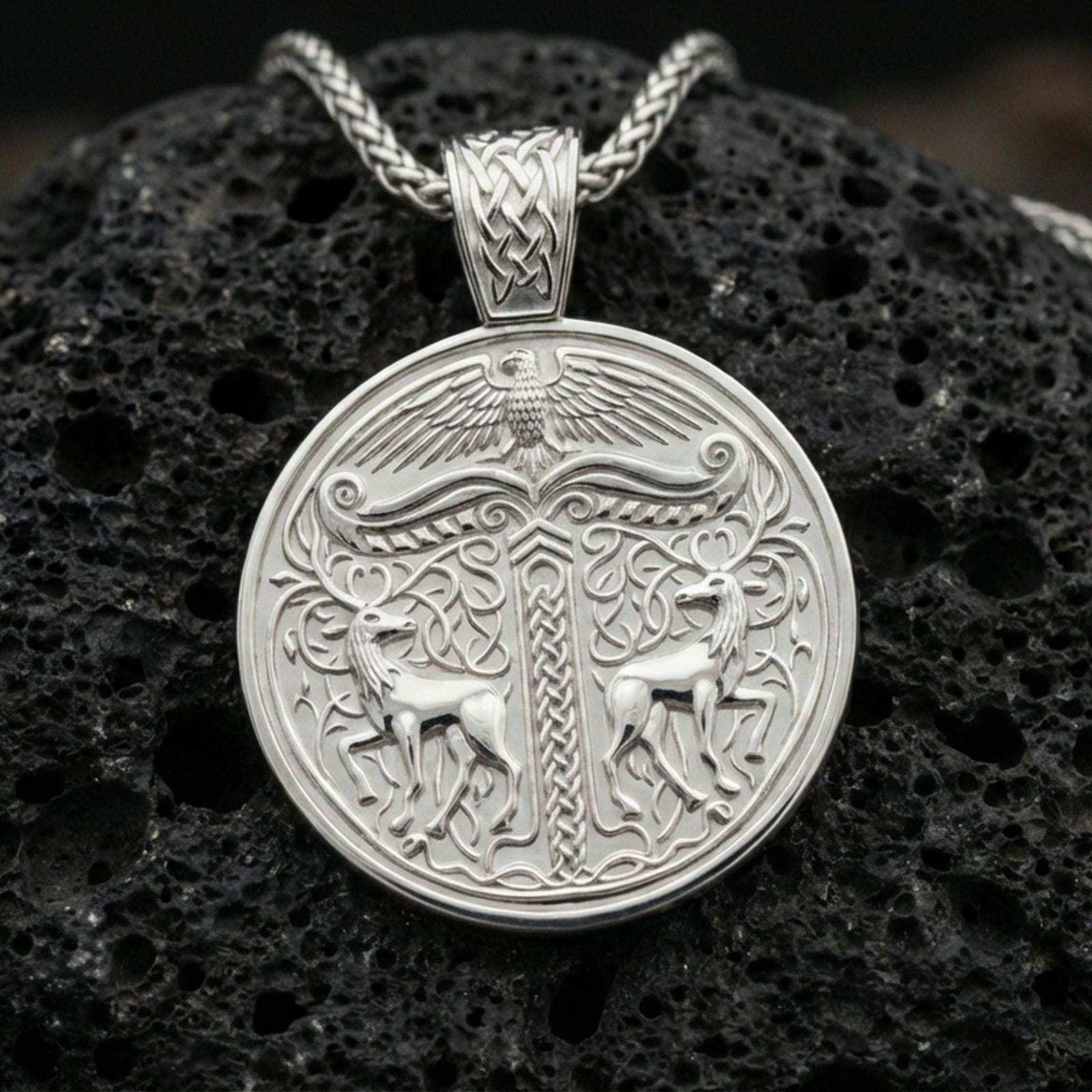 Polished silver Viking deer medallion pendant necklace on textured volcanic rock, crisp macro product photo emphasizing clean edges, sharp scroll engraving, and bright reflective highlights on the stags and border.