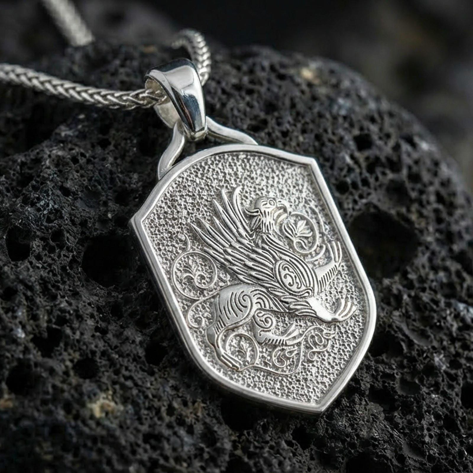 Polished silver griffin shield pendant necklace on textured volcanic rock, crisp macro product photo emphasizing clean edges, sharp feather lines, and bright reflective highlights on the raised relief.