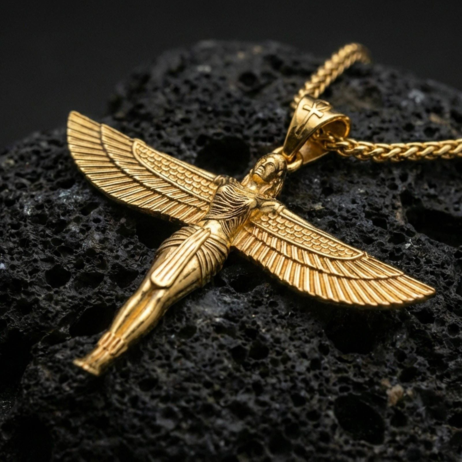 Gold plated winged Isis goddess pendant necklace on textured volcanic rock, premium close-up with warm reflective shine across the wings, body contours, and engraved textures.