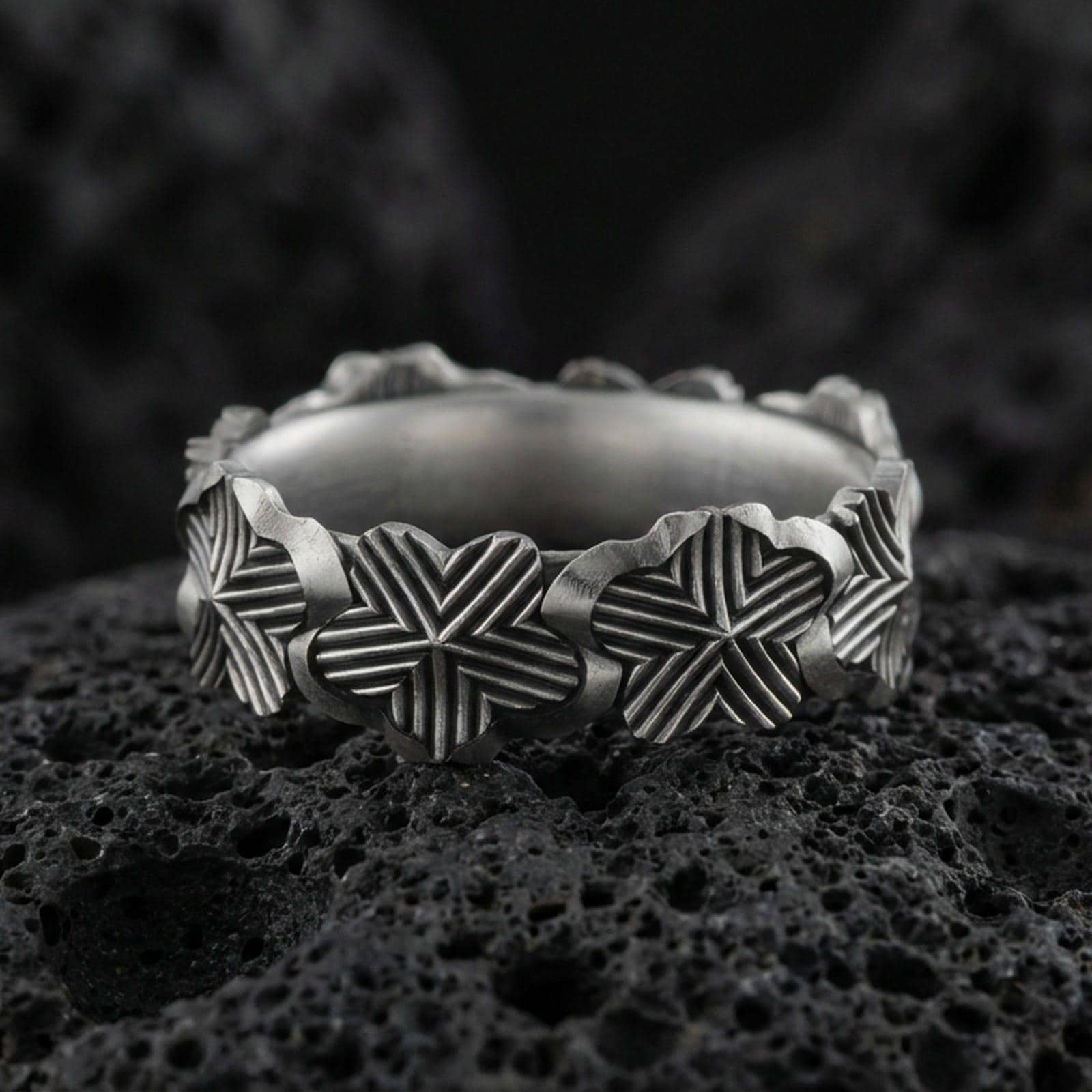 Oxidized floral band ring on textured volcanic rock, centered close-up highlighting the raised lily motifs, carved line texture, and deep antique shadowed contrast.
