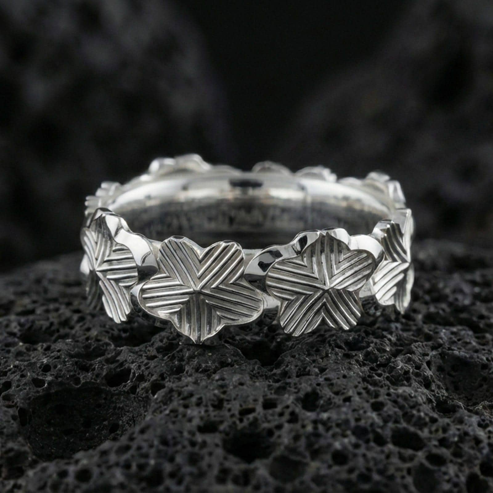 Polished silver floral band ring on textured volcanic rock, crisp macro product photo emphasizing clean edges, sharp flower relief, and bright reflective highlights across the patterned band.