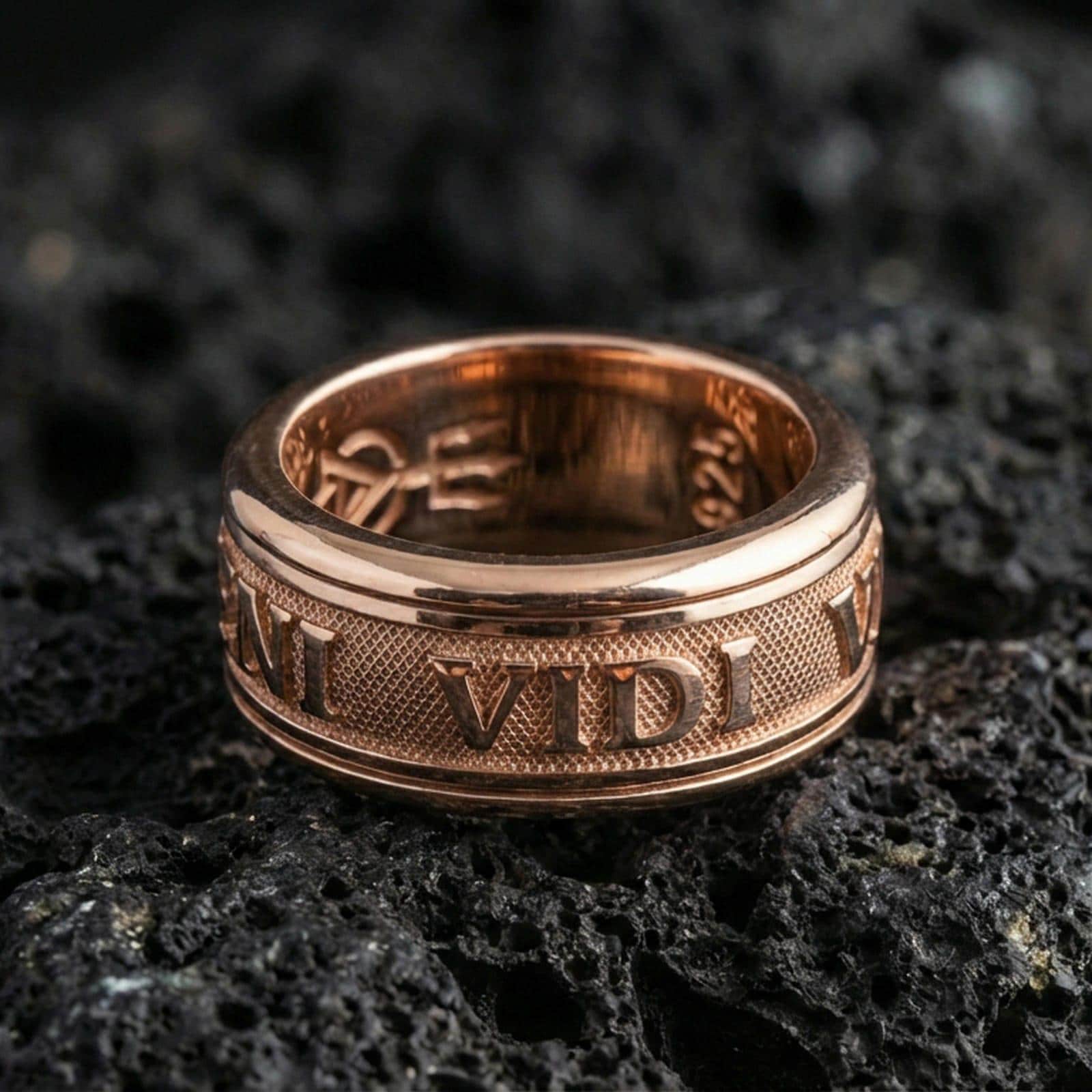 Rose gold plated Veni Vidi Vici spinner ring on textured volcanic rock, centered studio-style close-up showcasing soft rosy glow and crisp Latin quote engraving with a refined finish.
