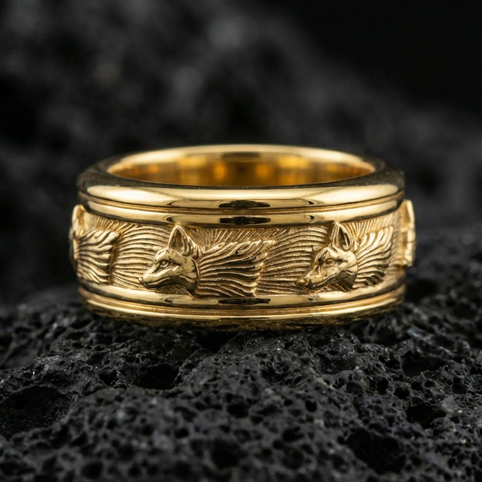 Gold plated wolf head band ring on textured volcanic rock, premium close-up with warm reflective shine across the wolf profiles, engraved fur lines, and raised ornamental contours.