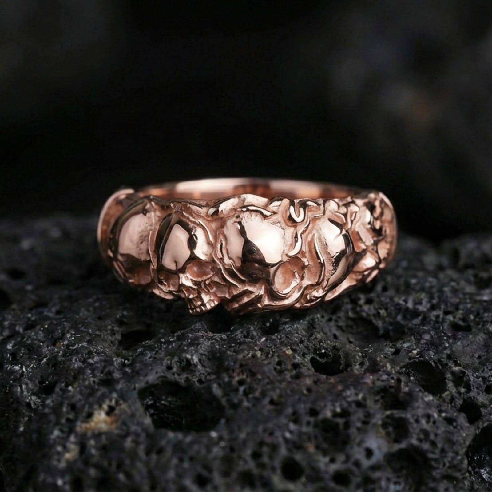 Rose gold plated skull band ring on textured volcanic rock, centered studio-style close-up showcasing soft rosy glow and crisp skull detailing with a refined finish.