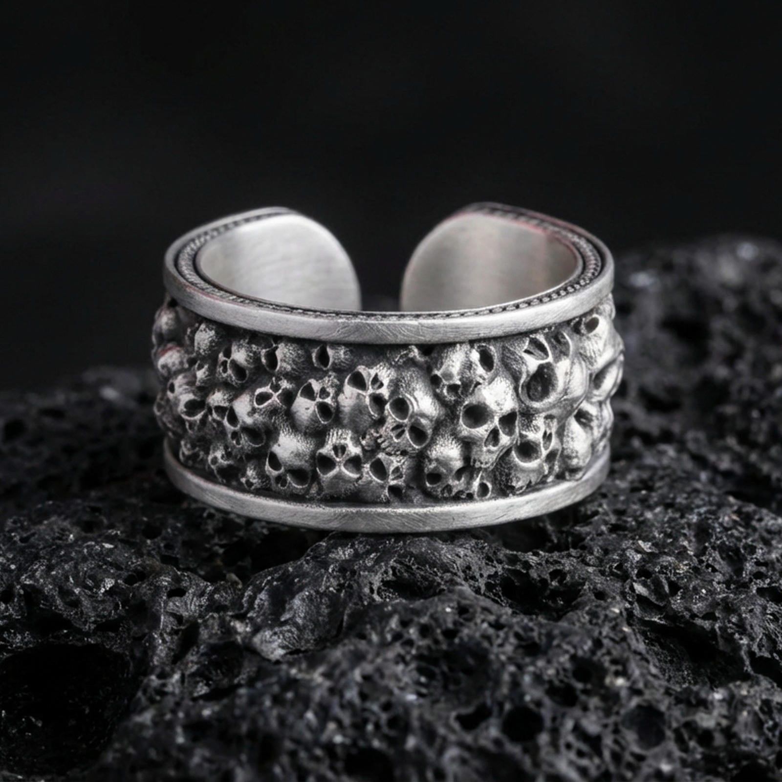 Oxidized skull cuff ring on textured volcanic rock, centered close-up highlighting the clustered skull texture, open band design, and deep antique shadowed contrast.