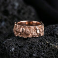 Load image into Gallery viewer, Rose gold plated skull and floral band ring on textured volcanic rock, centered studio-style close-up showcasing soft rosy glow and intricate skull-and-flower detailing with a refined finish.
