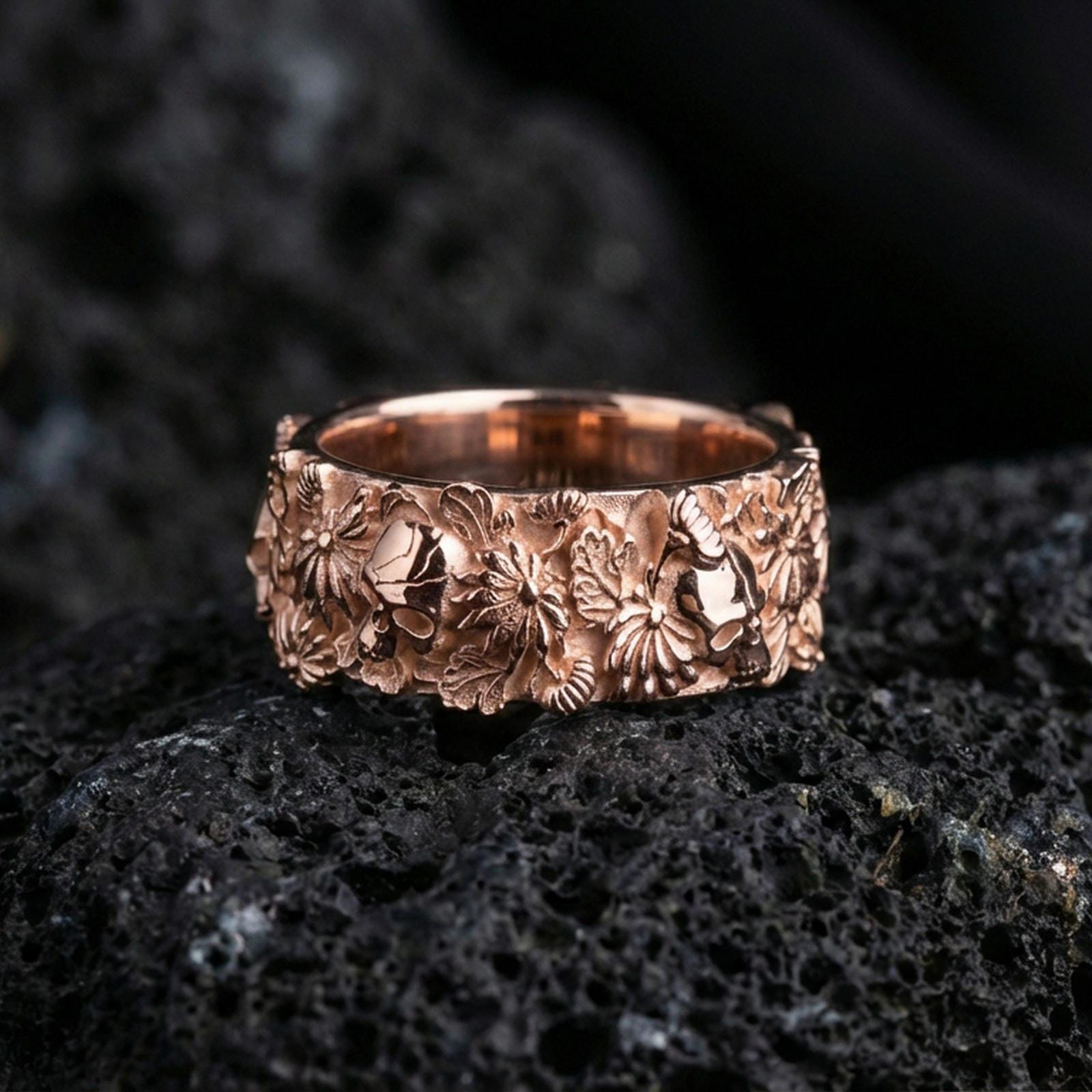 Rose gold plated skull and floral band ring on textured volcanic rock, centered studio-style close-up showcasing soft rosy glow and intricate skull-and-flower detailing with a refined finish.