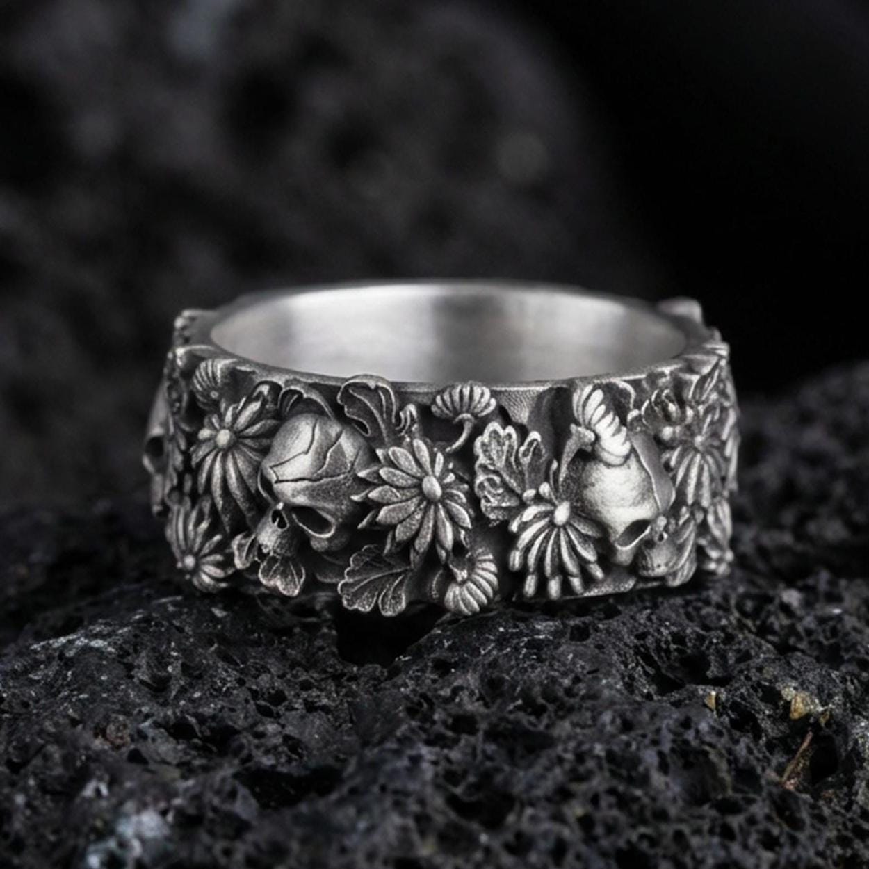 Oxidized skull and floral band ring on textured volcanic rock, centered close-up highlighting the raised skull accents, blooming flower relief, and deep antique shadowed contrast.
