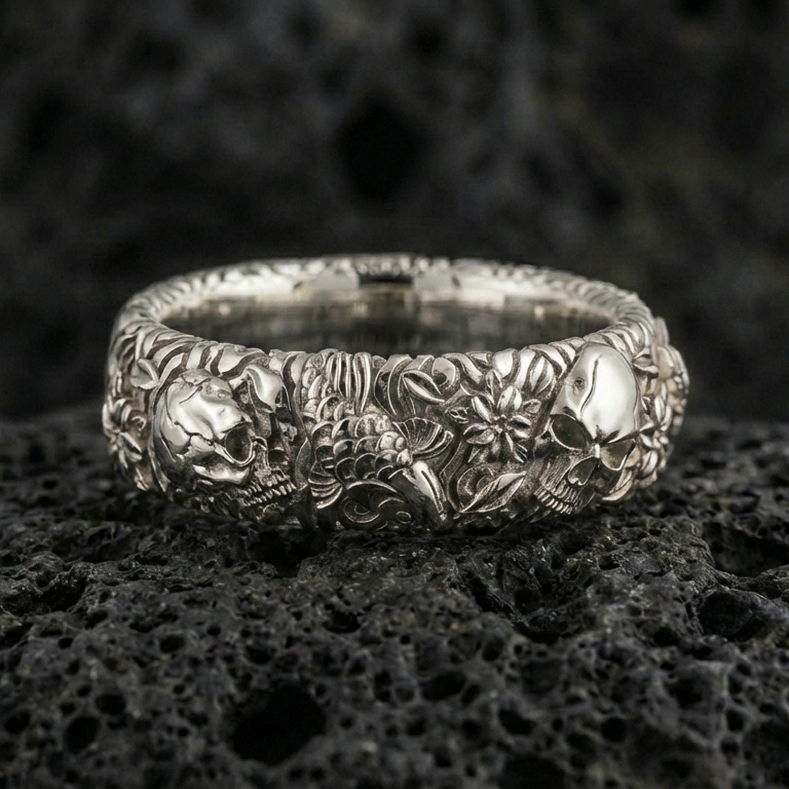 Polished silver skull and koi floral band ring on textured volcanic rock, crisp macro product photo emphasizing clean edges, sharp koi-and-flower engraving, and bright reflective highlights across the sculpted band.
