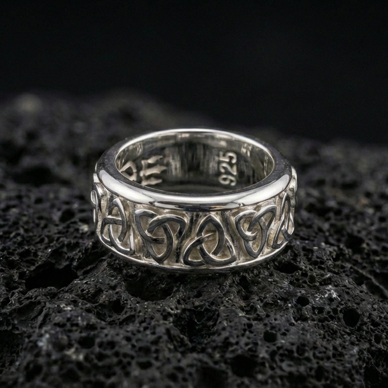 Polished silver Celtic triquetra band ring on textured volcanic rock, crisp macro product photo emphasizing clean edges, sharp knotwork relief, and bright reflective highlights across the band.