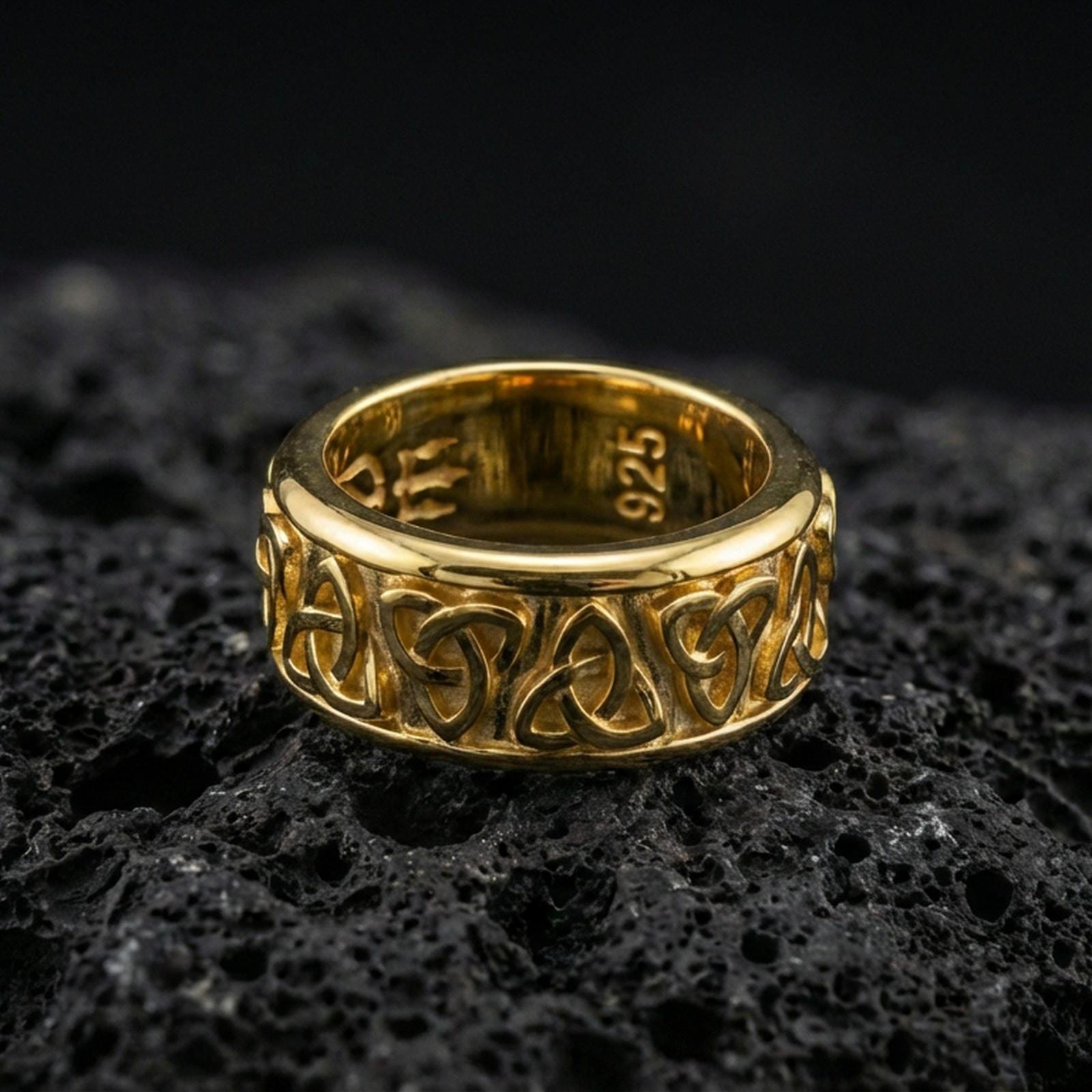 Gold plated Celtic triquetra band ring on textured volcanic rock, premium close-up with warm reflective shine across the repeating Trinity knots and raised contours.