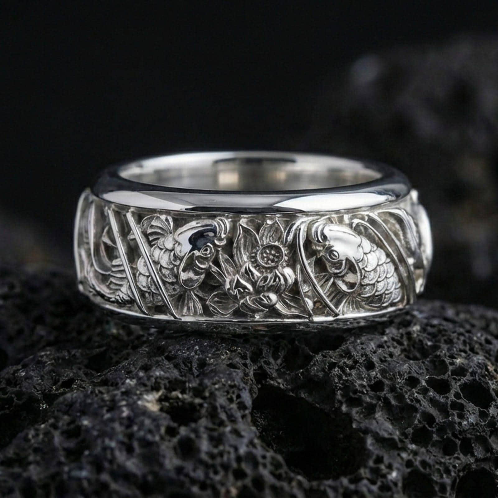 Polished silver koi fish and lotus band ring on textured volcanic rock, crisp macro product photo emphasizing clean edges, sharp scale engraving, and bright reflective highlights across the koi-and-lotus details.