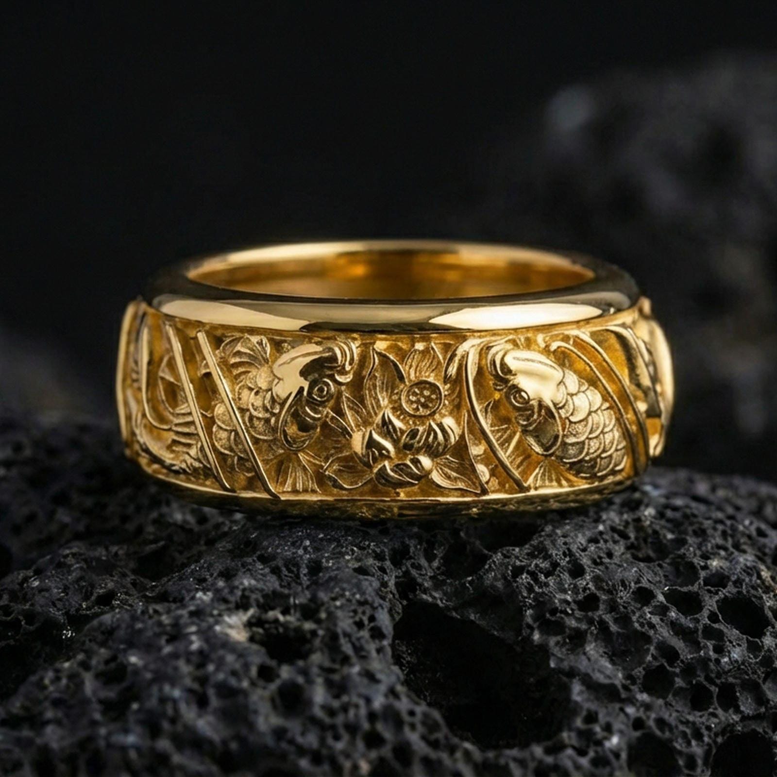 Gold plated koi fish and lotus band ring on textured volcanic rock, premium close-up with warm reflective shine across the koi motifs, lotus petals, and raised ornamental contours.