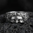 Load image into Gallery viewer, Oxidized face relief band ring on textured volcanic rock, centered close-up highlighting the sculpted eyes and flowing hair detail with deep antique shadowed contrast.
