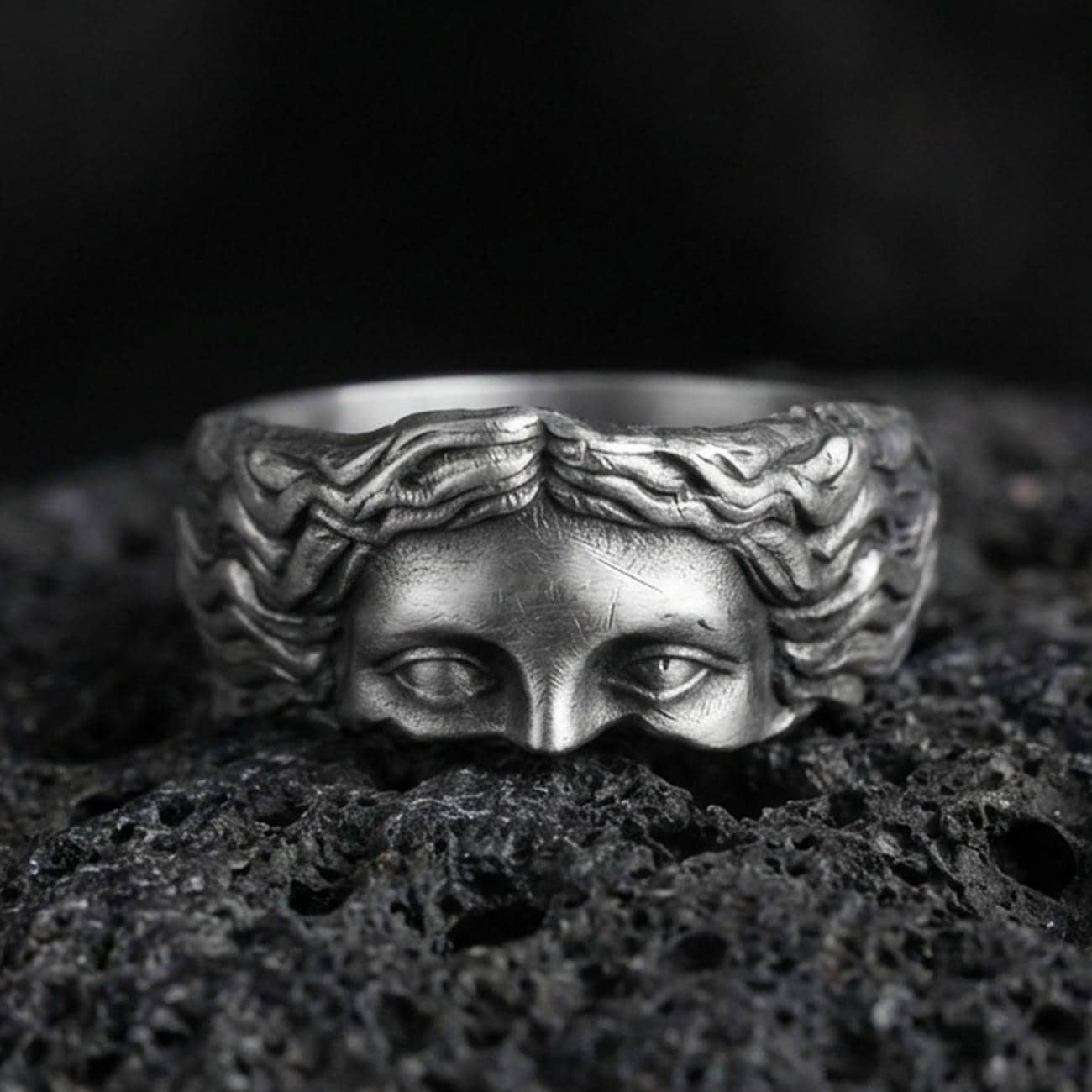 Oxidized face relief band ring on textured volcanic rock, centered close-up highlighting the sculpted eyes and flowing hair detail with deep antique shadowed contrast.