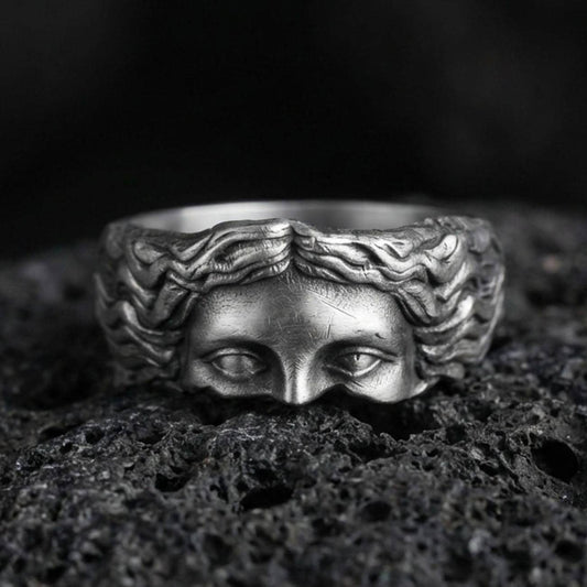 Oxidized face relief band ring on textured volcanic rock, centered close-up highlighting the sculpted eyes and flowing hair detail with deep antique shadowed contrast.
