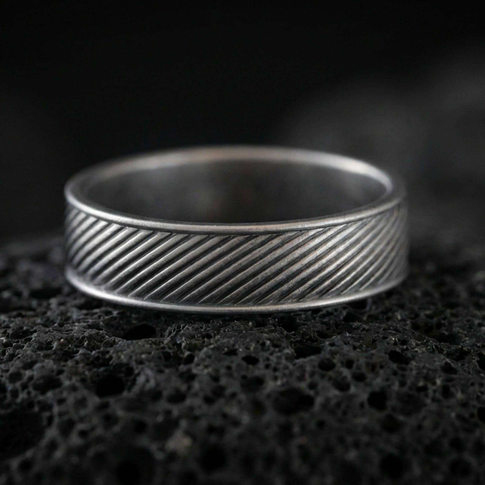 Oxidized diagonal groove band ring on textured volcanic rock, centered close-up highlighting the slanted line engraving and deep antique shadowed contrast across the channel pattern.
