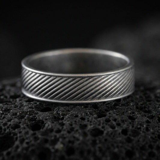Oxidized diagonal groove band ring on textured volcanic rock, centered close-up highlighting the slanted line engraving and deep antique shadowed contrast across the channel pattern.