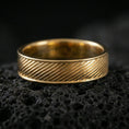 Load image into Gallery viewer, Gold plated diagonal groove band ring on textured volcanic rock, premium close-up with warm reflective shine across the slanted groove texture and smooth rounded edges.
