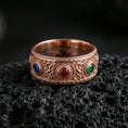 Load image into Gallery viewer, Rose gold plated ornate gemstone band ring on textured volcanic rock, refined macro product shot capturing soft rosy highlights on the ornate band with the blue, red, and green stones clearly visible.
