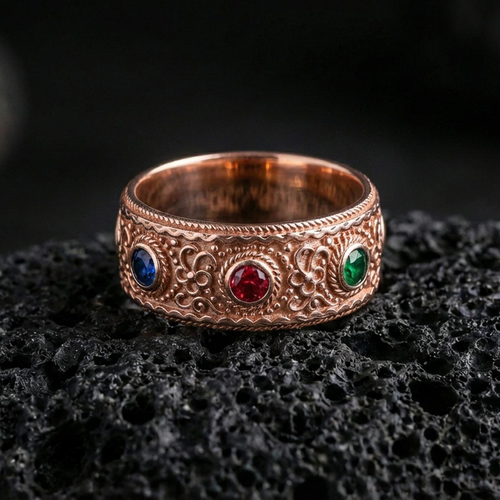 Rose gold plated ornate gemstone band ring on textured volcanic rock, refined macro product shot capturing soft rosy highlights on the ornate band with the blue, red, and green stones clearly visible.