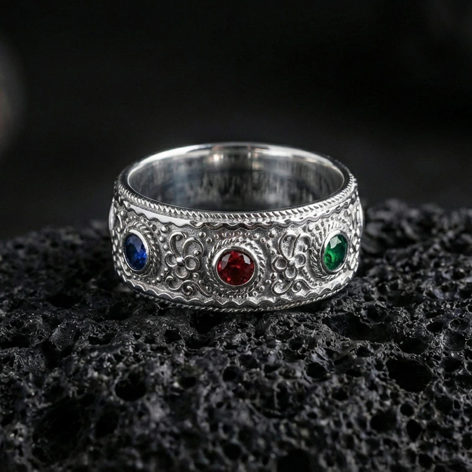 Polished silver ornate gemstone band ring on textured volcanic rock, crisp studio macro emphasizing the raised scrollwork, beaded borders, and three gemstone settings with bright reflective shine.
