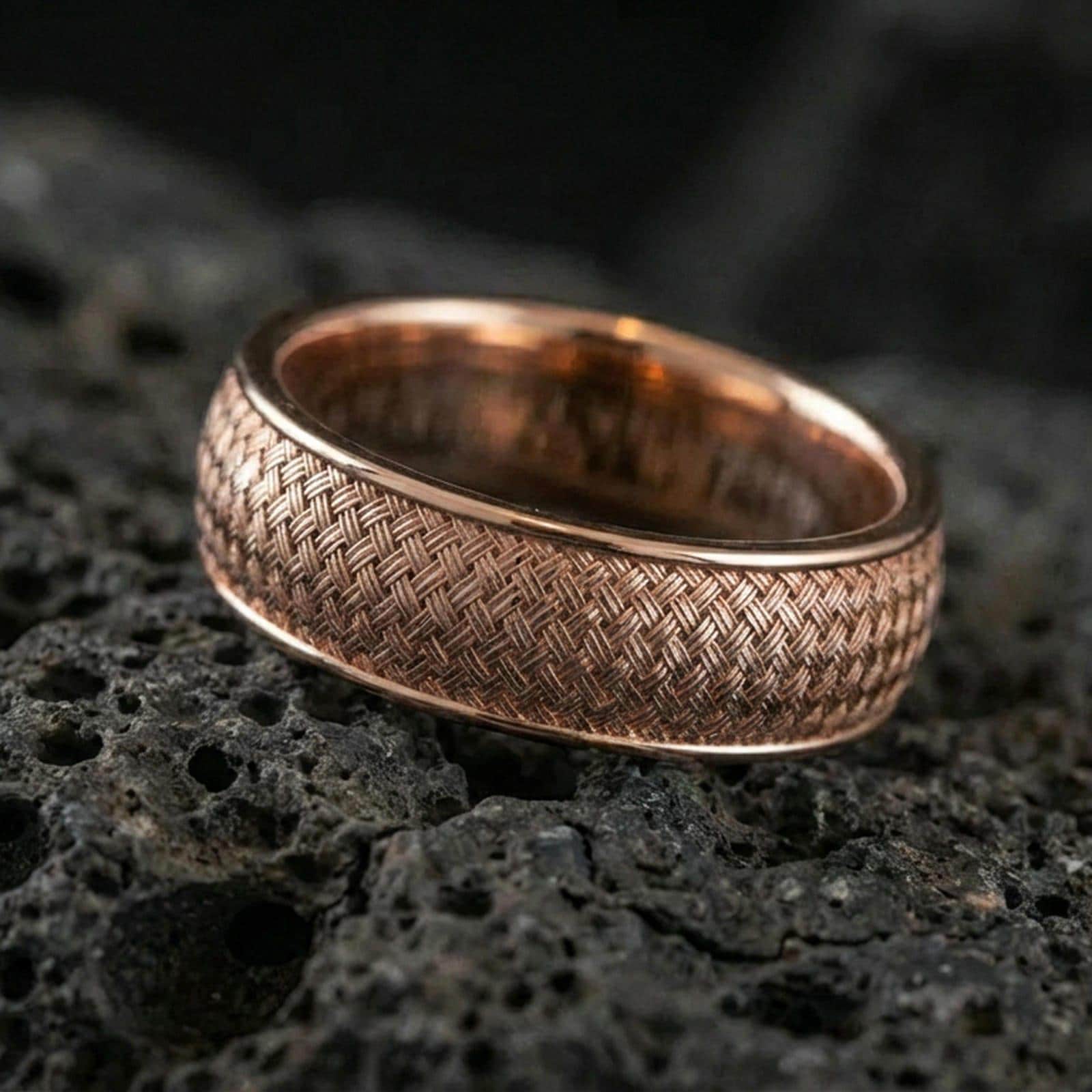 Rose gold plated woven texture band ring on textured volcanic rock, refined macro product shot capturing soft rosy highlights on the woven pattern and rounded band profile.