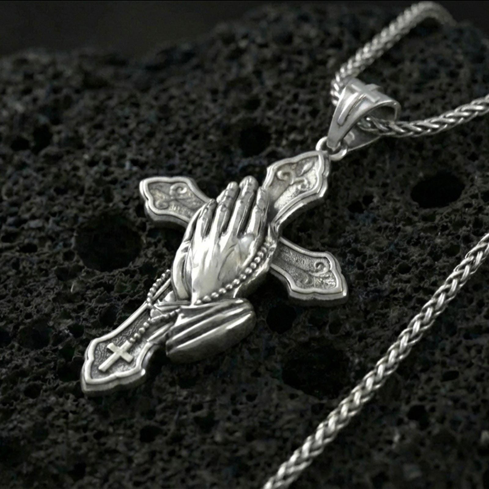 Polished silver praying hands cross pendant on porous volcanic stone, close-up highlighting the raised hand sculpture, crisp cross edges, and clean reflective finish