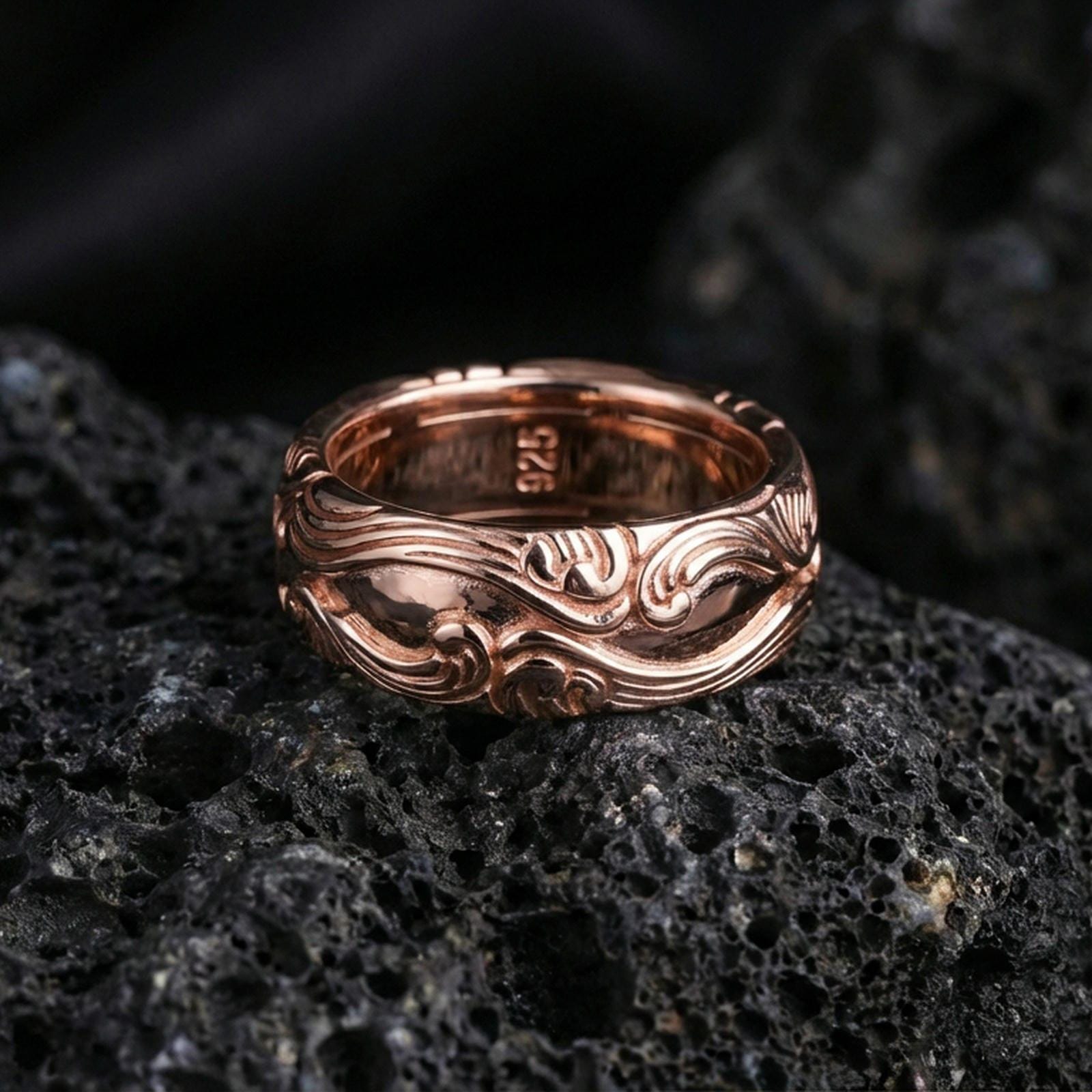 Rose gold plated scrollwork band ring on textured volcanic rock, refined macro product shot capturing soft rosy highlights and the dimensional scroll engraving.