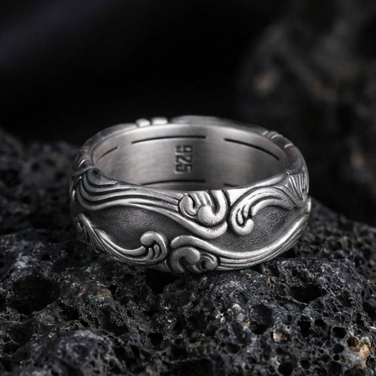 Oxidized scrollwork band ring on textured volcanic rock, macro close-up highlighting the darkened recessed background and raised flowing wave motifs.