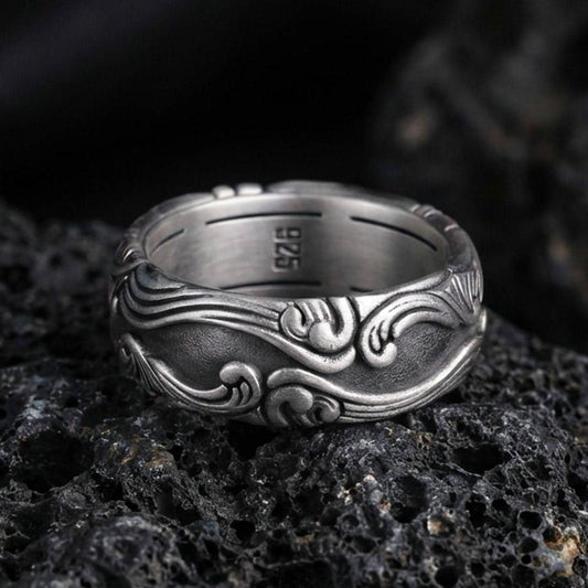 Oxidized scrollwork band ring on textured volcanic rock, macro close-up highlighting the darkened recessed background and raised flowing wave motifs.