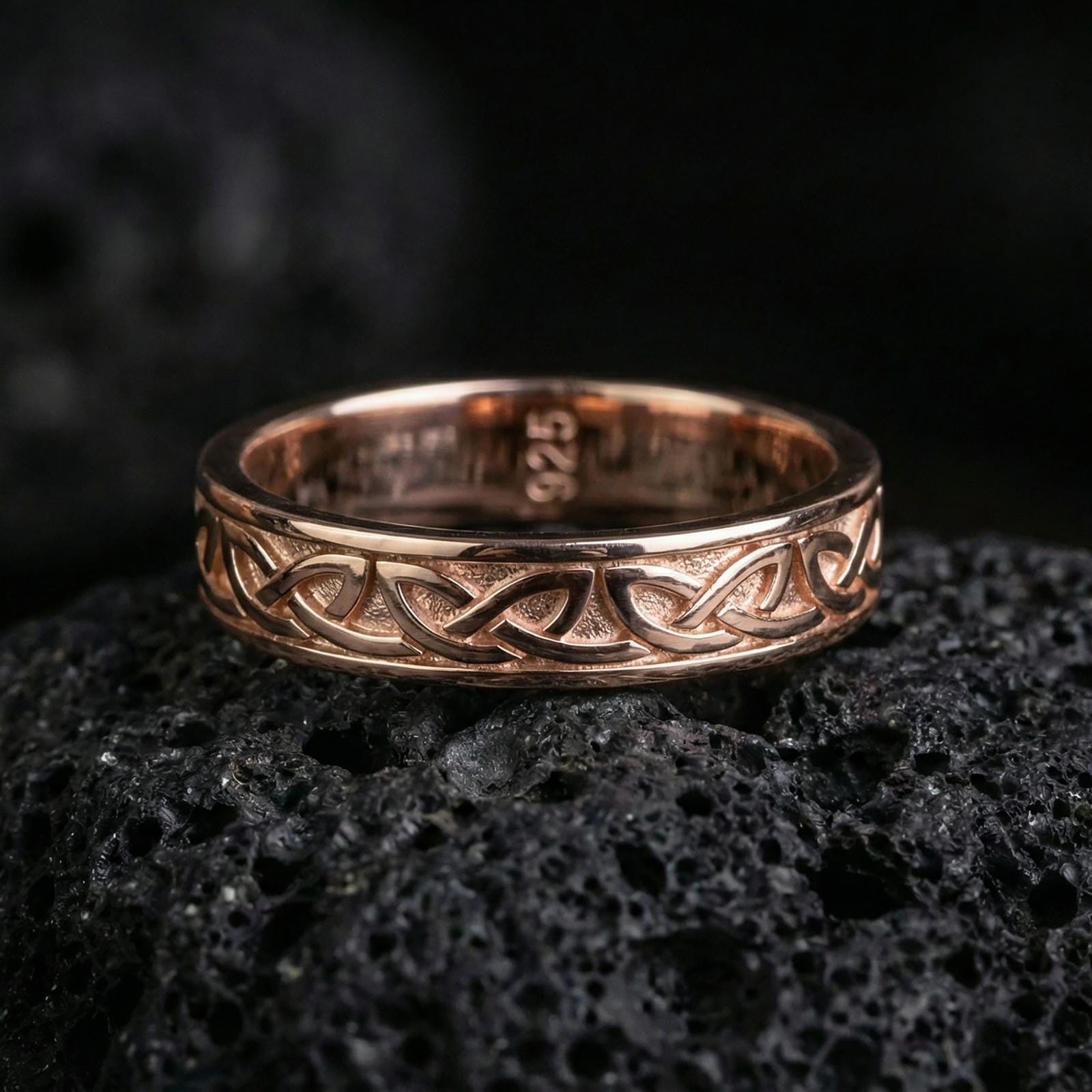 Rose gold plated Celtic knot band ring on volcanic stone, refined macro shot with soft rosy highlights and detailed interlaced knot engraving.