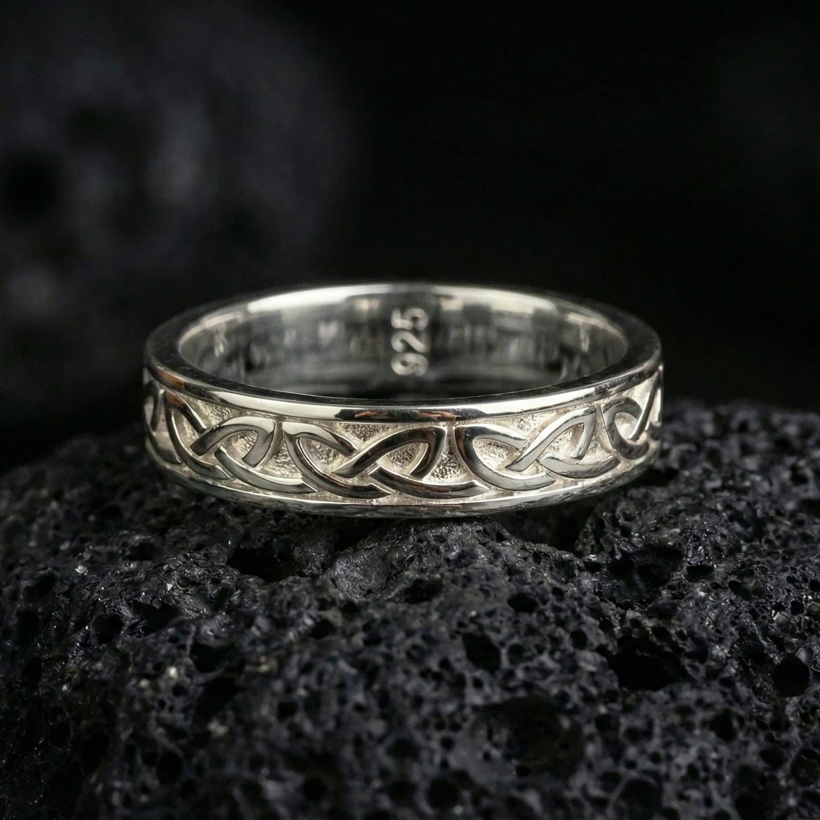 Polished silver Celtic knot band ring on volcanic stone, sharp macro product photo emphasizing clean reflective edges and crisp knotwork relief.