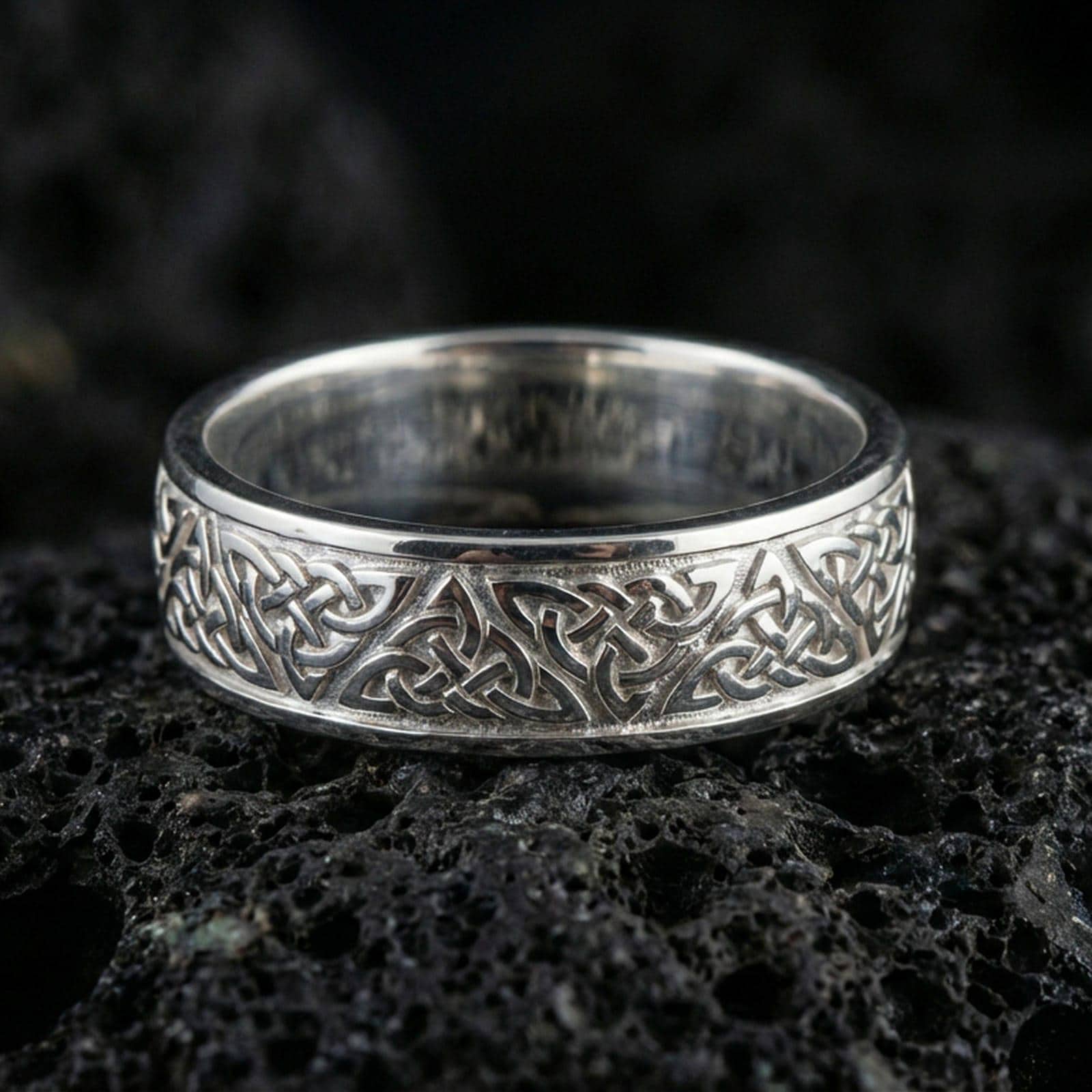 Polished silver Celtic knot band ring on volcanic stone, crisp macro product photo emphasizing clean reflective edges and sharp engraving depth.