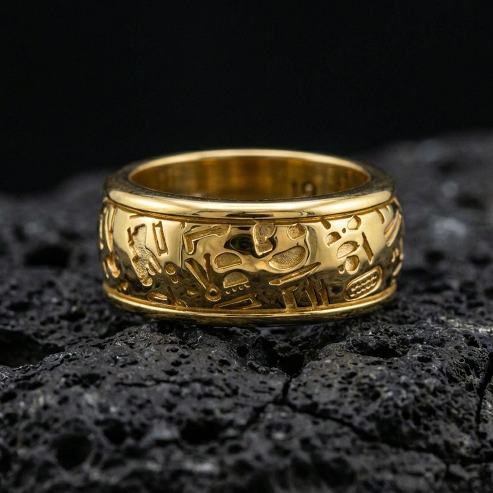 Gold plated hieroglyph band ring on volcanic stone, premium close-up showing warm gold tone over the carved Egyptian hieroglyph pattern.